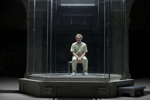 A man is held in a giant glass prison cell dressed in a green jumpsuit