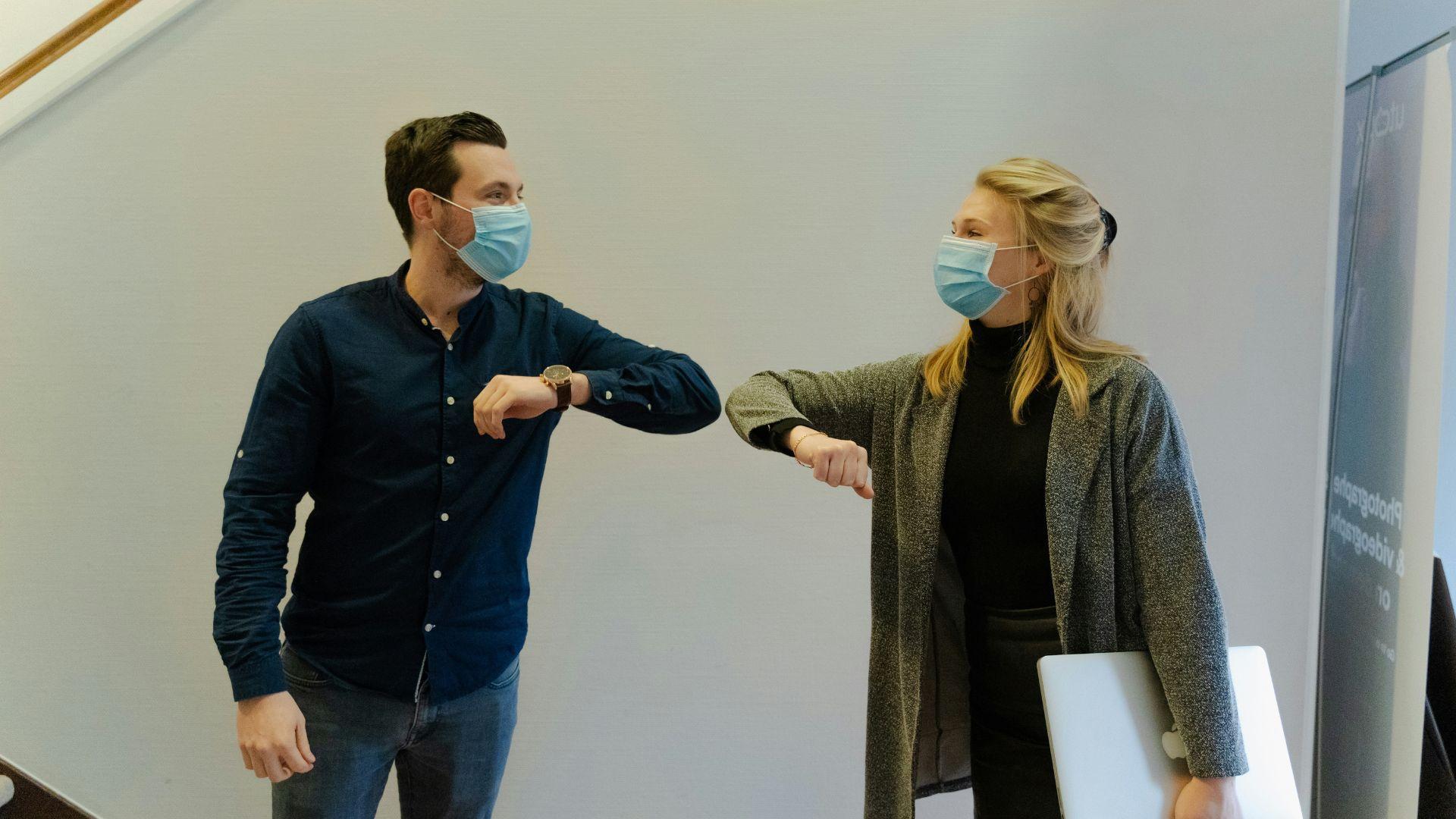 A man and a woman, both wearing surgical face masks, greet each other by touching elbows in an indoor setting. The man is dressed in a dark blue shirt and jeans, and the woman is wearing a grey coat over a black outfit