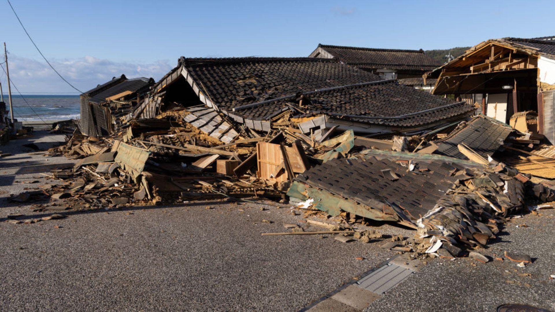 Collapsed traditional houses with dark tiled roofs lie in ruins after a disaster. Debris is scattered across the street in the foreground, with the ocean visible in the background under a clear blue sky