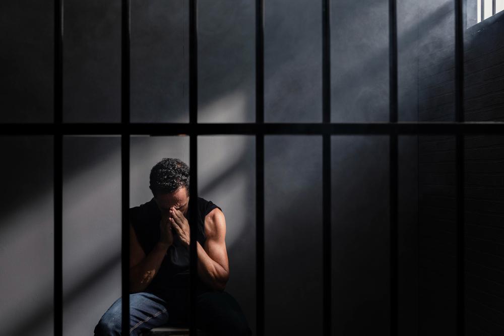 A man dressed in a black sleeveless shirt holds his face in his hands behind bars in prison