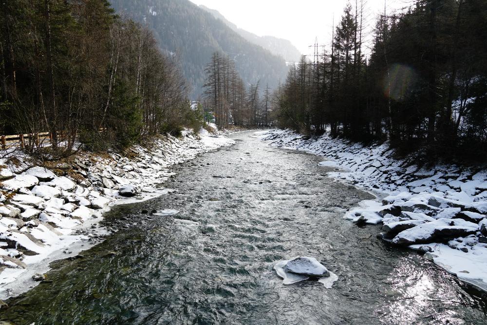 A giant river slows down a mountainous region flanked on either side by snow-covered forests