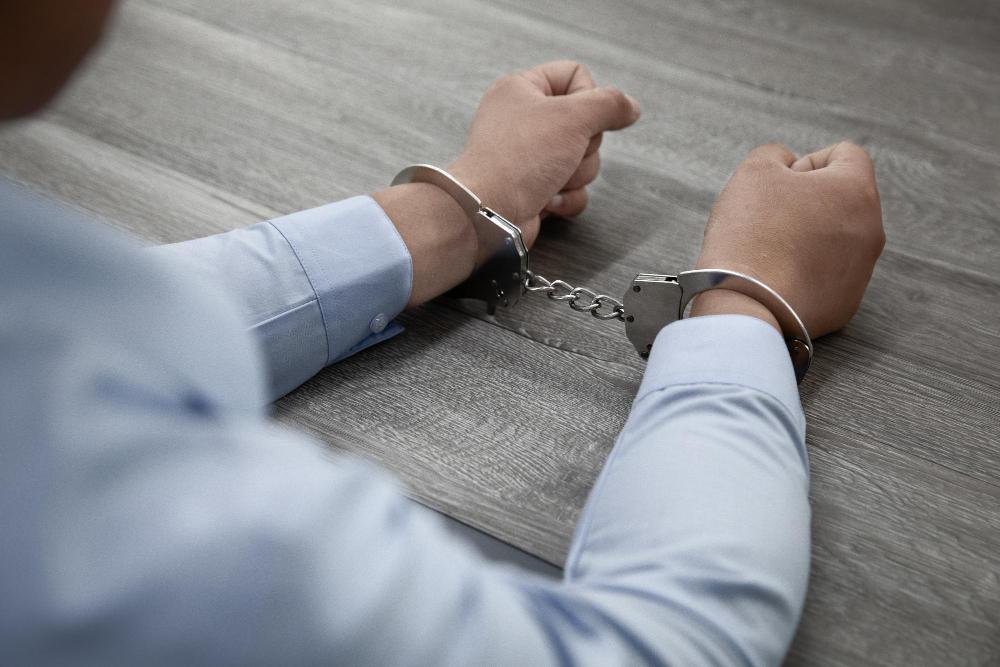 A man dressed in a light blue shirt sits at a wooden table with his hands in cuffs