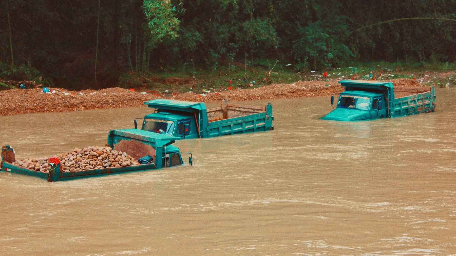 Two teal trucks are submerged in a muddy flood, with the closest one carrying a load of rocks. The water level reaches the trucks' cabins, suggesting a severe flood event