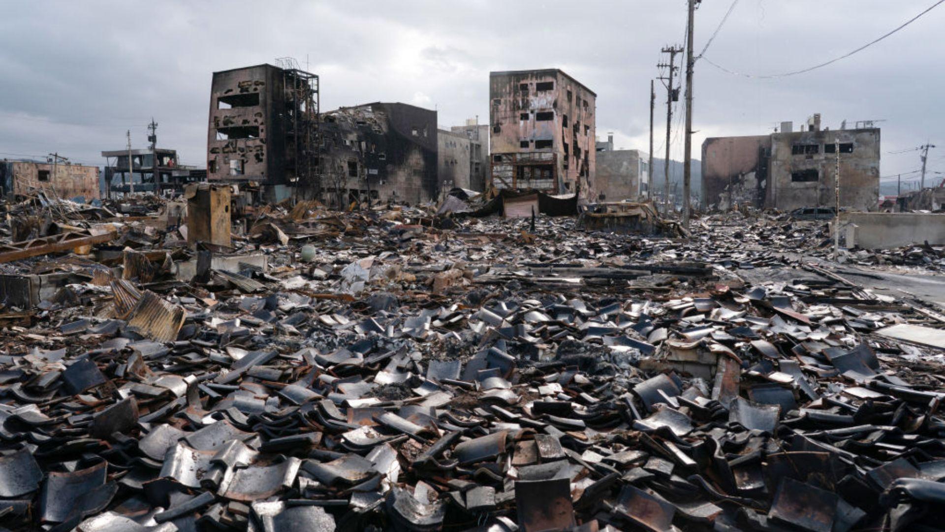 An image depicting the ruins of a devastated urban area. Charred remains of buildings with hollowed-out windows stand in the background. The foreground is covered with a vast spread of debris,