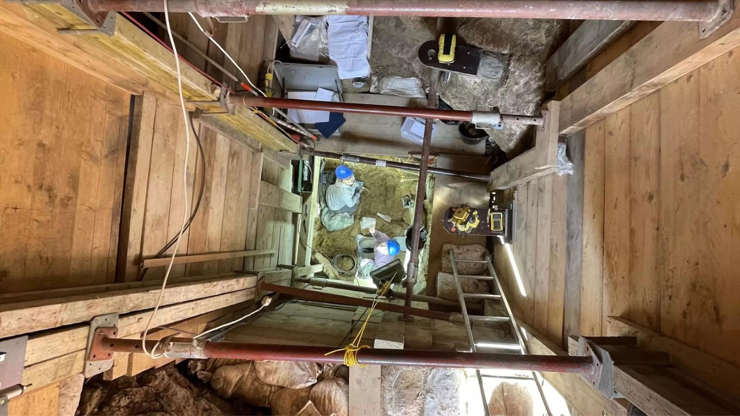 An overhead view of an archaeological dig site. The image shows a deep, rectangular pit reinforced with wooden planks and metal beams. At the bottom of the pit, several archaeologists in hard hats are working, surrounded by tools and equipment, including shovels, buckets, and measuring instruments