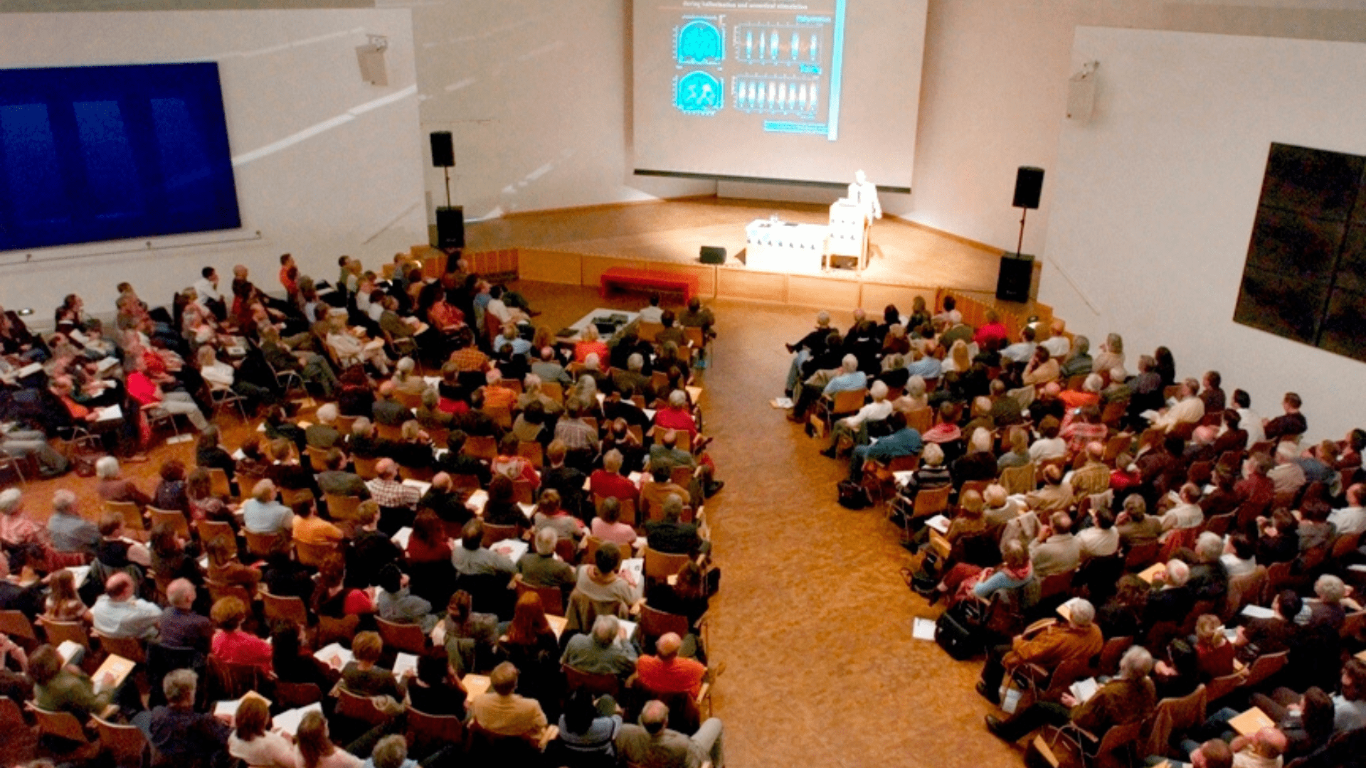 A lecture hall filled with people listening to someone giving a presentation on stage.