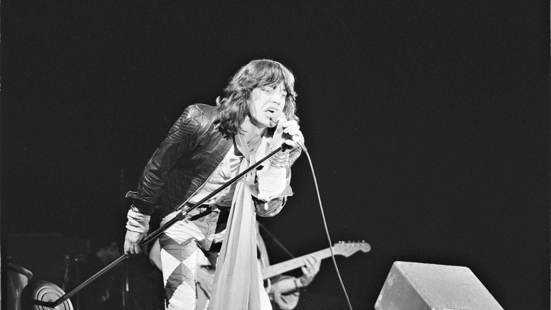 A black and white image of Mick Jagger leaning into a microphone stand, passionately singing or shouting during a performance. He is dressed in a leather jacket and patterned shirt