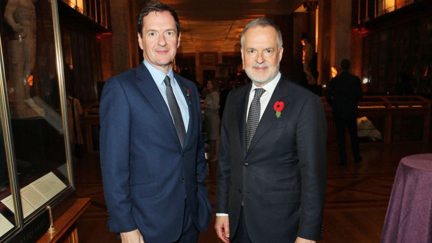 Two men dressed in business suits, one in blue and the other in gray, stand side by side posing for a photograph in a museum gallery. Both men are wearing red poppy pins on their lapels