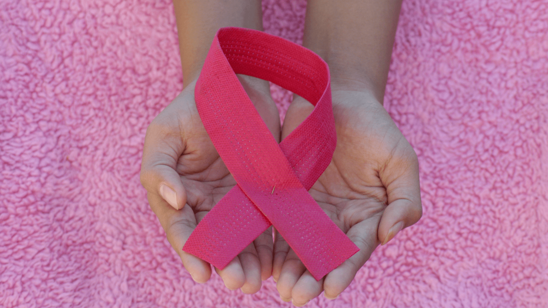 A person holding a pink breast cancer ribbon above a pink fabric surface.
