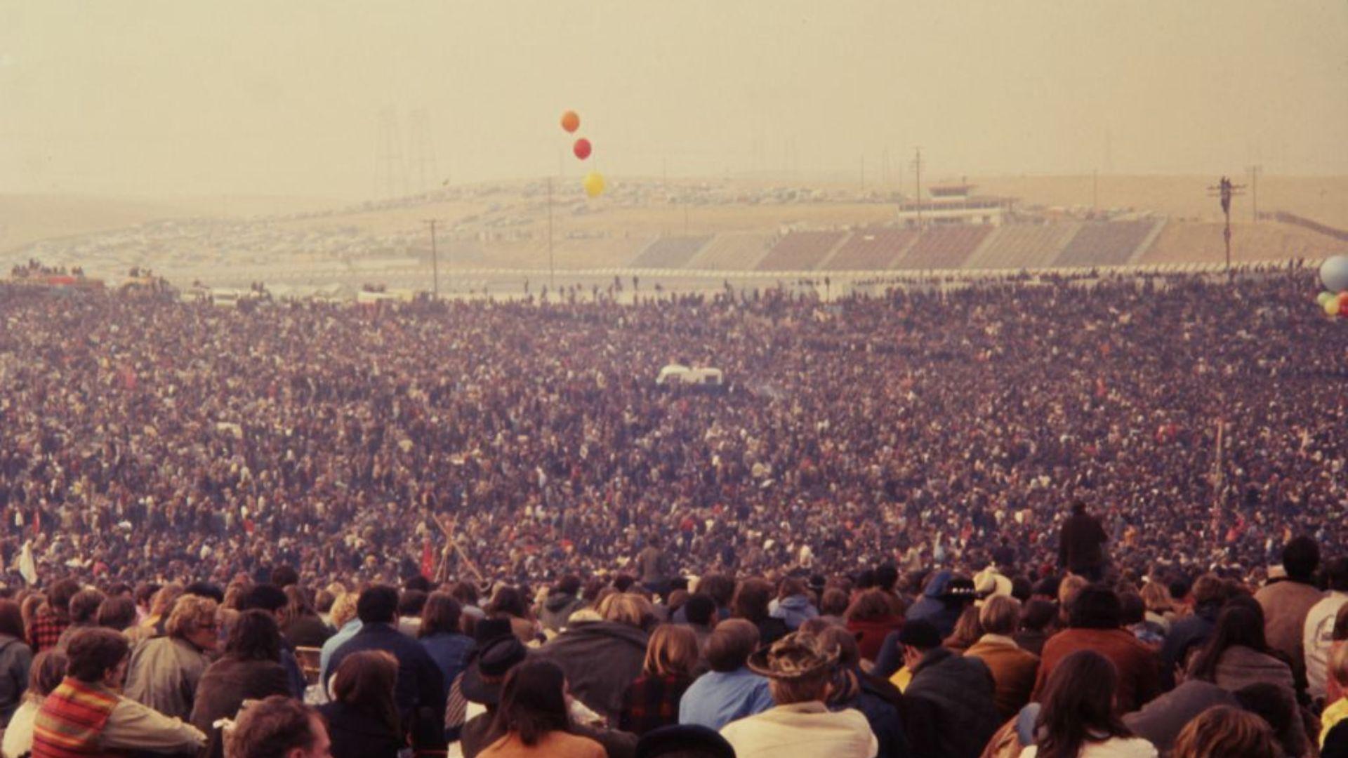 A vast sea of people covers the landscape at an outdoor concert venue, with the crowd stretching back to the distant horizon. In the foreground, spectators are seated, looking towards the stage area, which is not visible in the photo