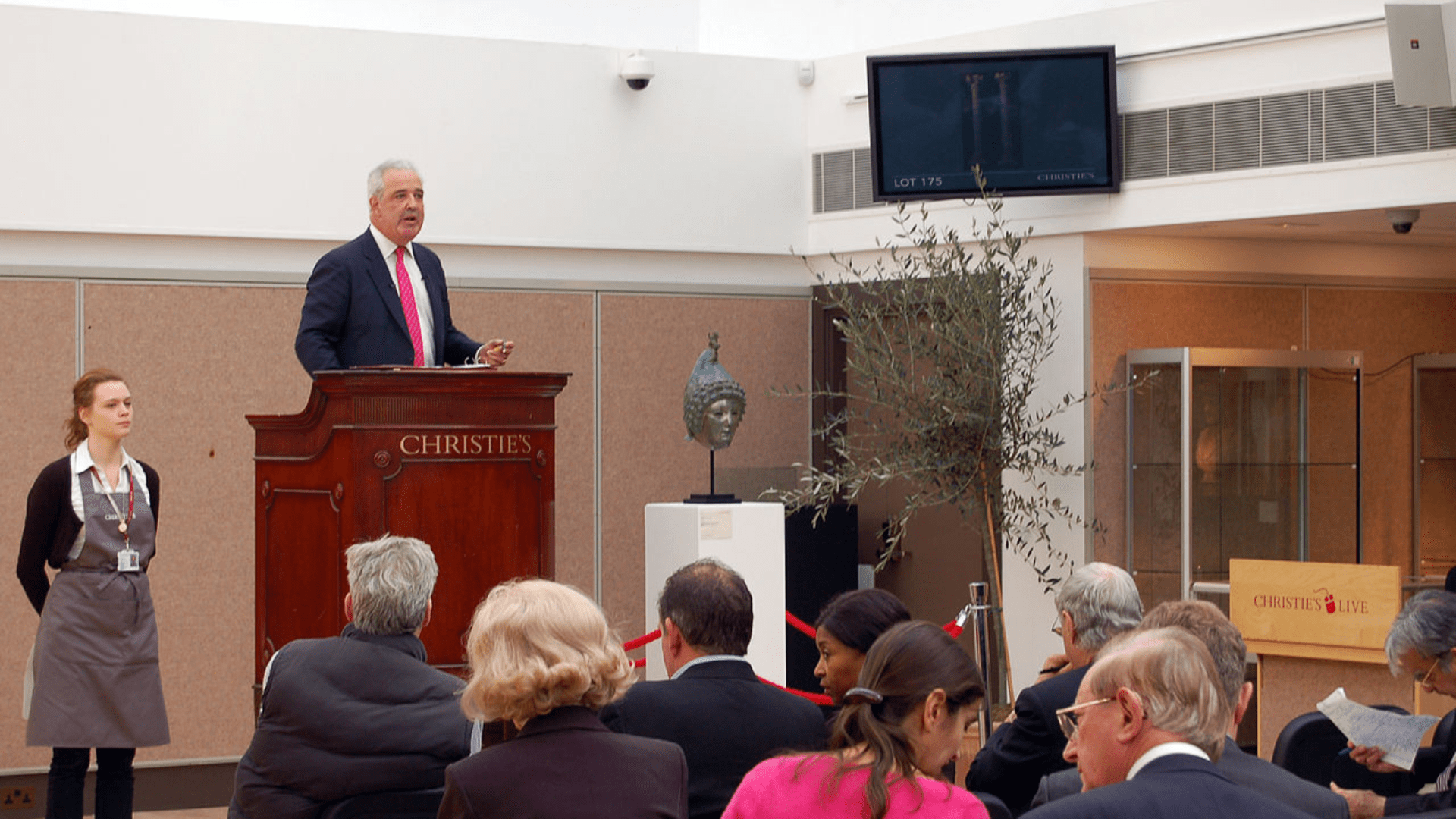 An auctioneer stands at a podium in front of a crowd of auction-goers. A helmet is on display next to the auctioneer’s podium.