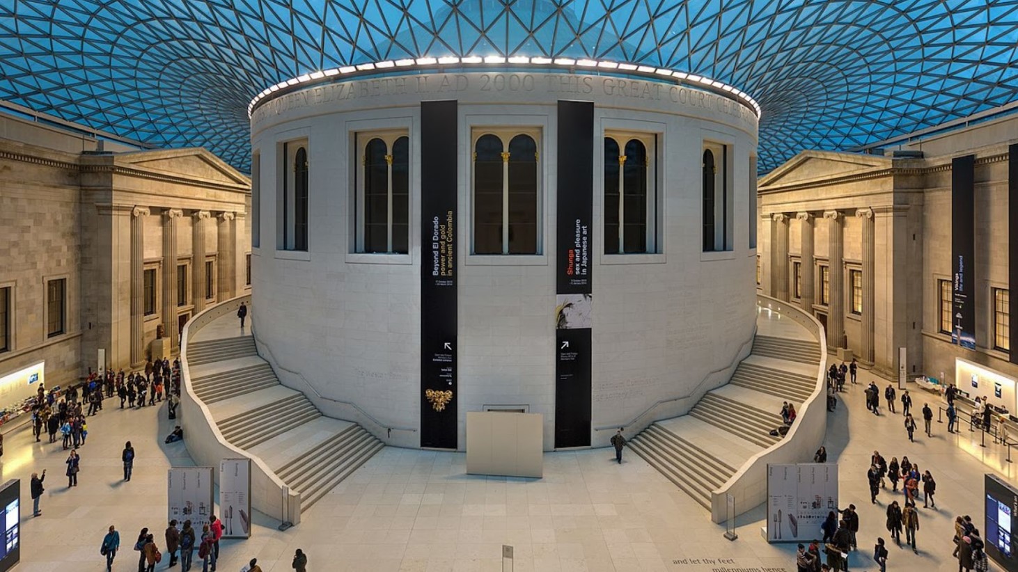 A panoramic view of the Great Court within the British Museum, showcasing the expansive space with a geometric glass roof. Visitors are scattered around the court