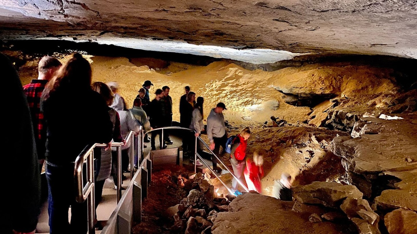 A group of people walking on a metal walkway through a large, well-lit cavern. The cave ceiling looms overhead