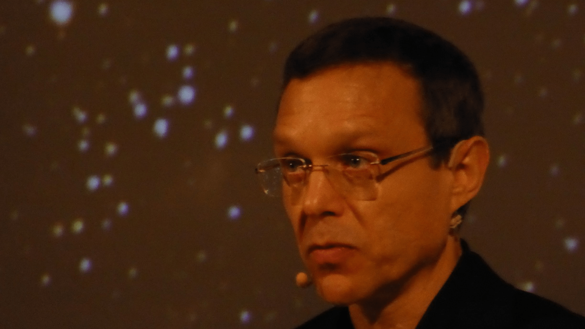 A photo of Dr. Avi Loeb’s head and neck at an angle, with a background of blurry specks.