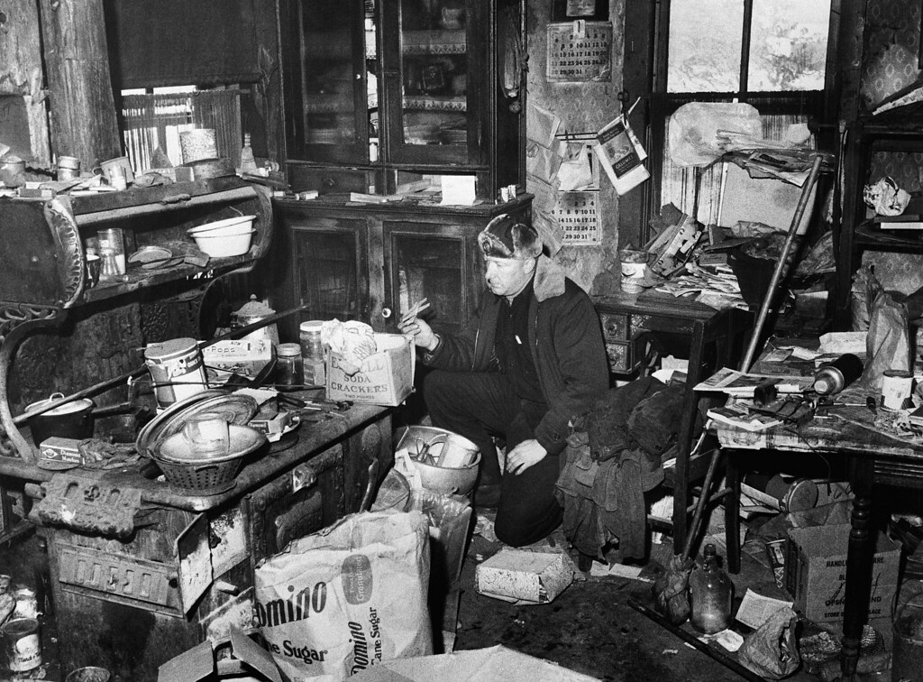Unidentified police officer examines the junk-littered kitchen in the farm home of Edward Gein, where authorities found human skulls and other parts of human bodies. They also found the butchered body of Mrs. Bernice Worden hung in a shed near the house.