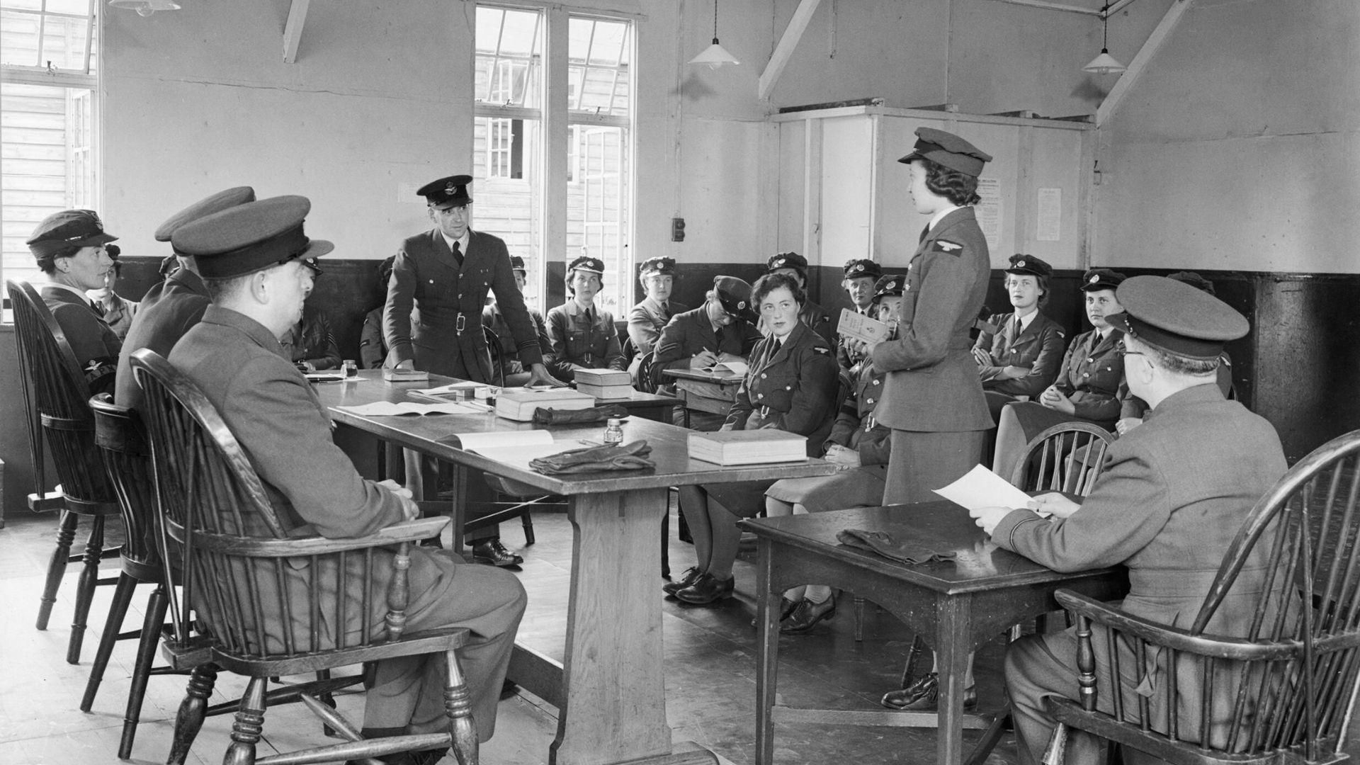 A black and white photo of women and men during World War II in a room talking, depicting a meeting of the Women's Auxiliary Air Force.