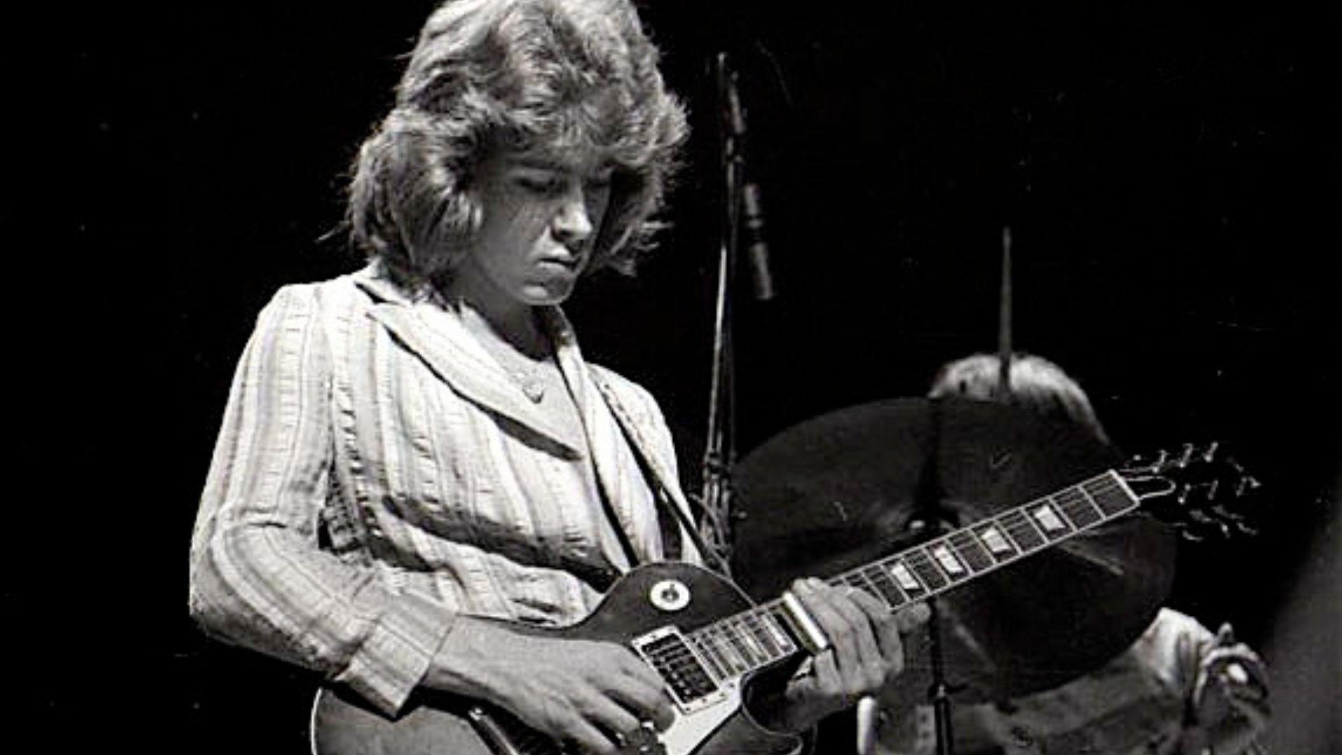 A black and white image of Mick Taylor onstage playing slide guitar