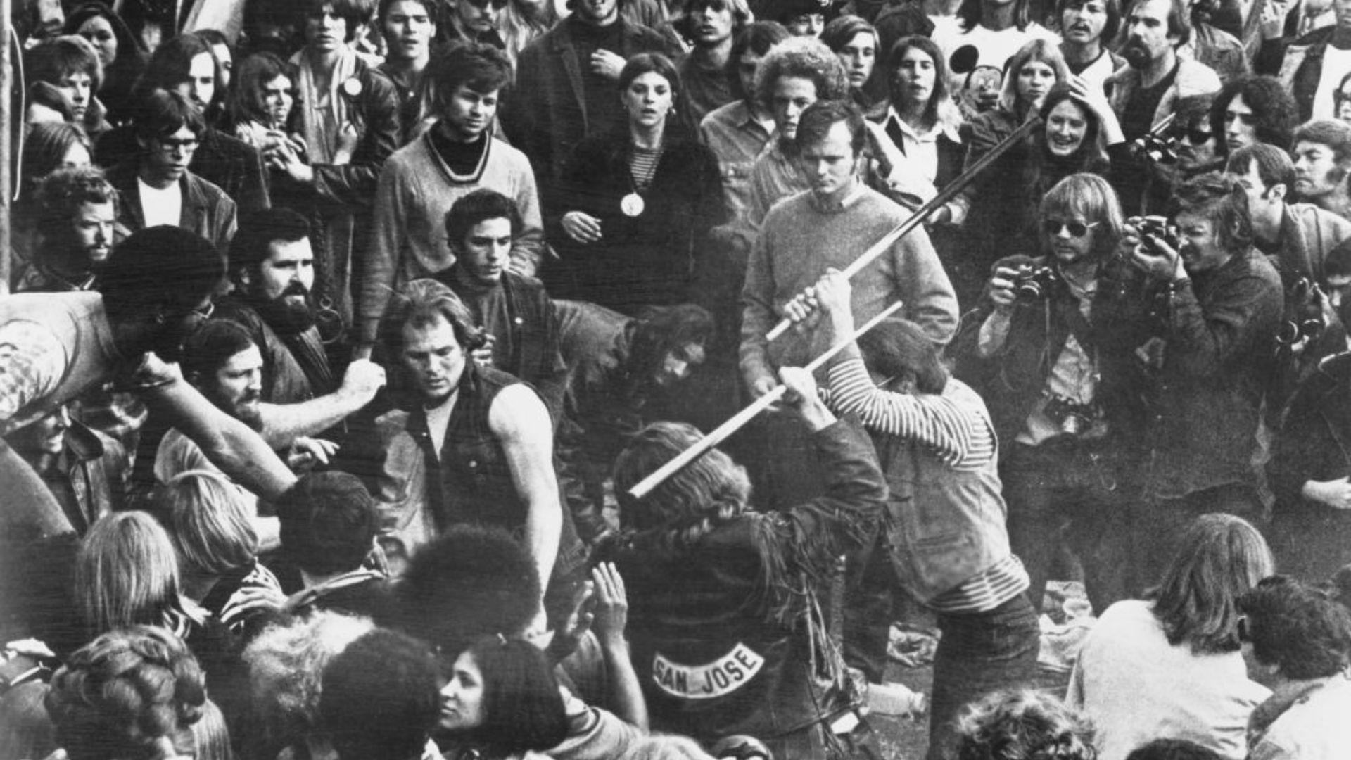 A black and white photo capturing a tense moment within a dense crowd at a concert. Several individuals appear to be engaged in a physical altercation, with one person wielding a pool cue. Onlookers have varied expressions of concern and shock. A person with a 'San Jose' patch on their jacket is visible in the foreground