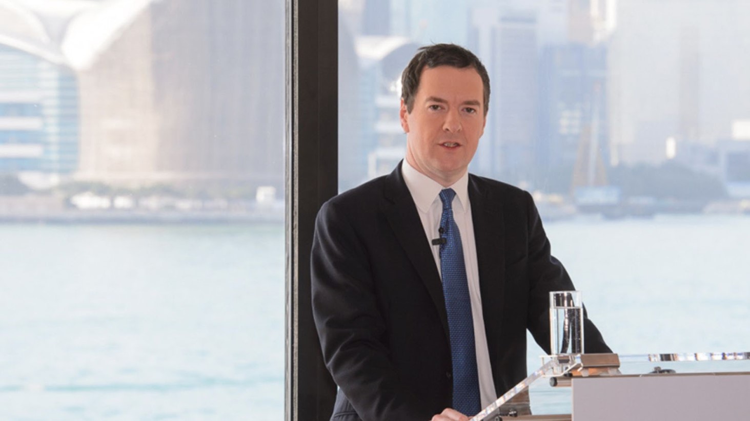 George Osborne in a dark suit and blue tie stands behind a lectern with an open briefcase, presenting to an audience with a blurred city skyline and water visible through the window behind him
