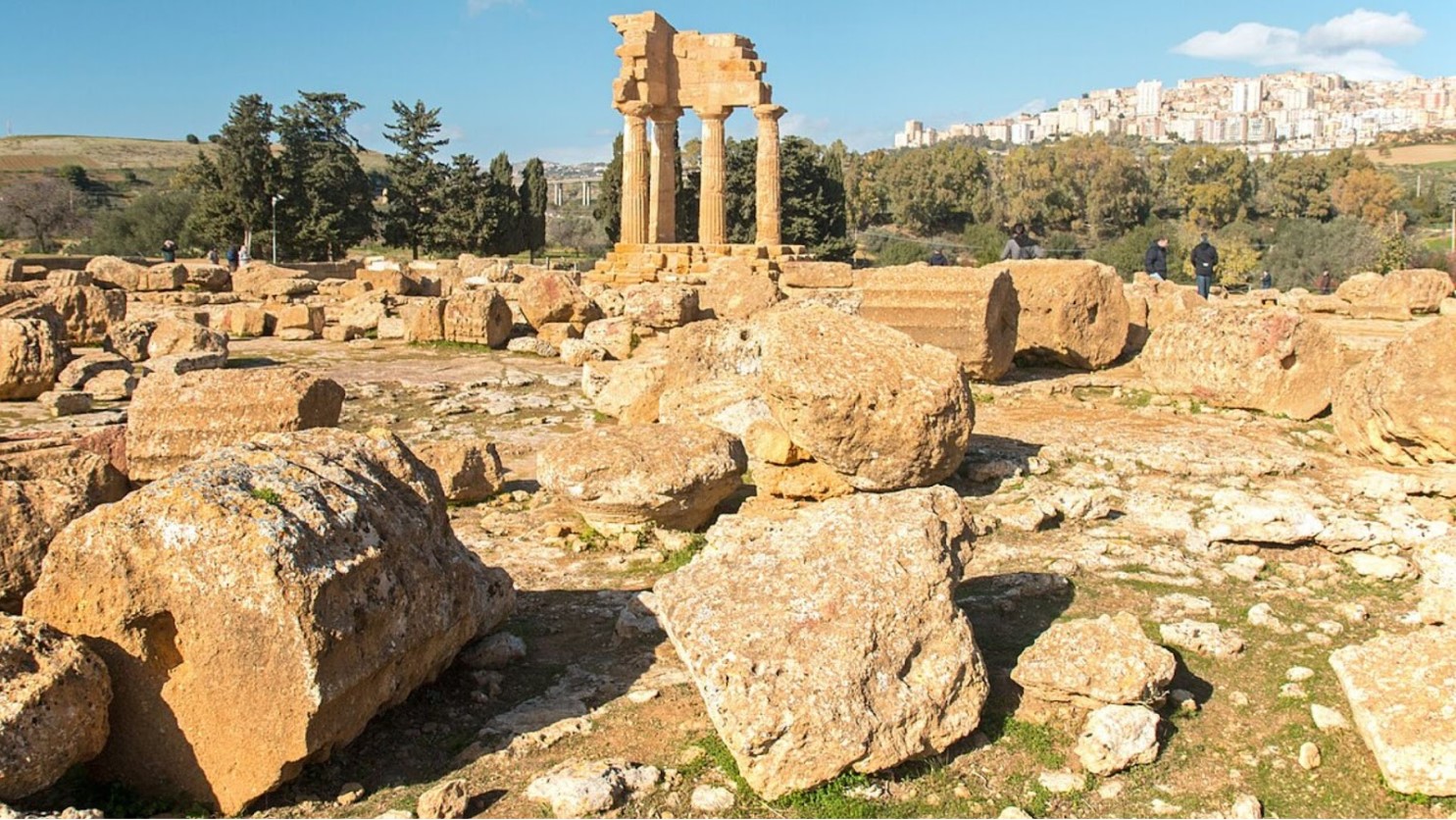 The ruined remains of Greek columns cover the ground with several left standing. In the background, a modern city on a hill.