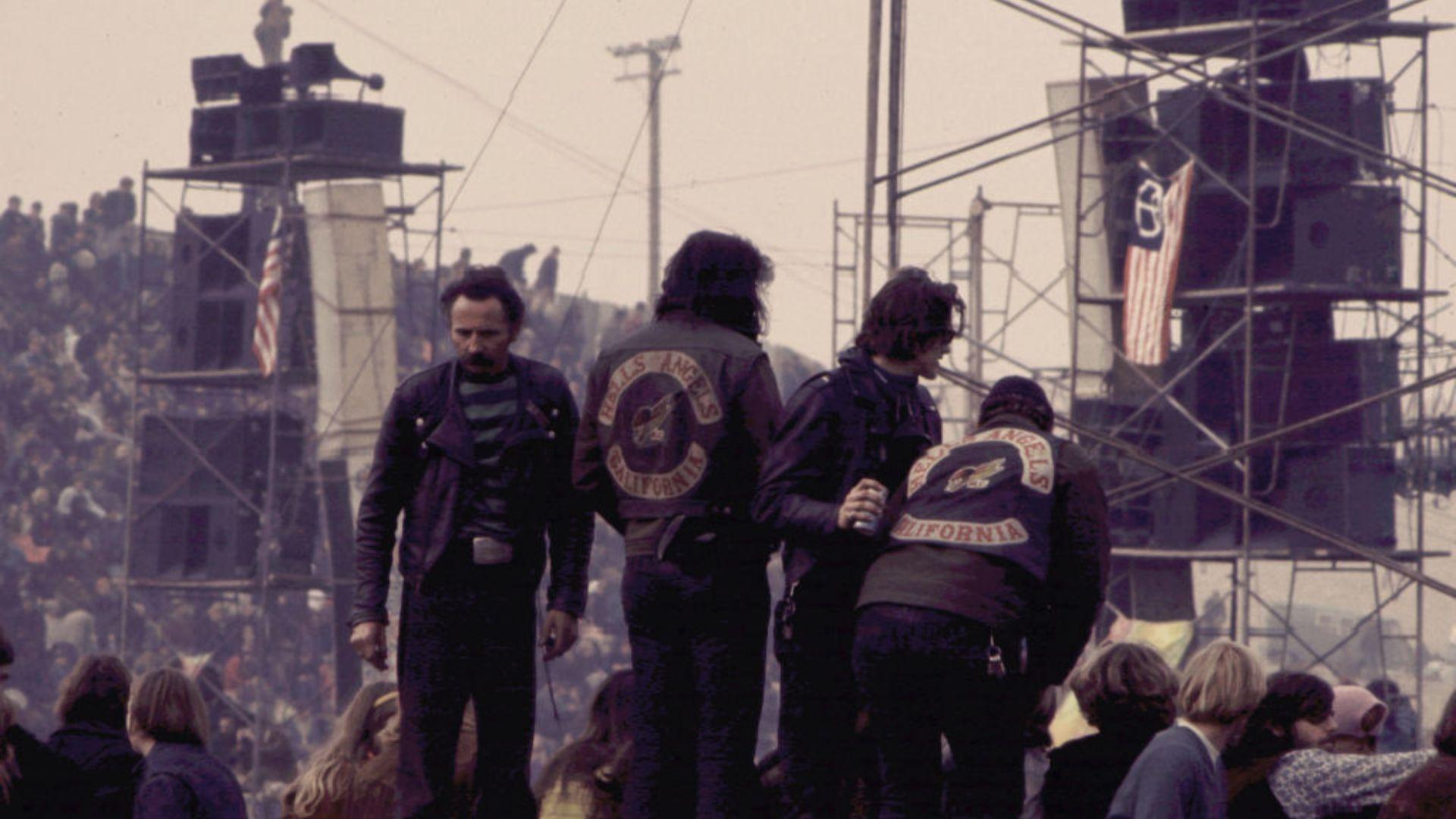 Three individuals wearing jackets with the 'Hells Angels California' emblem on the back are standing amidst a crowd at an outdoor event. In the background, a large number of people can be seen sitting on scaffolding structures beside an American flag