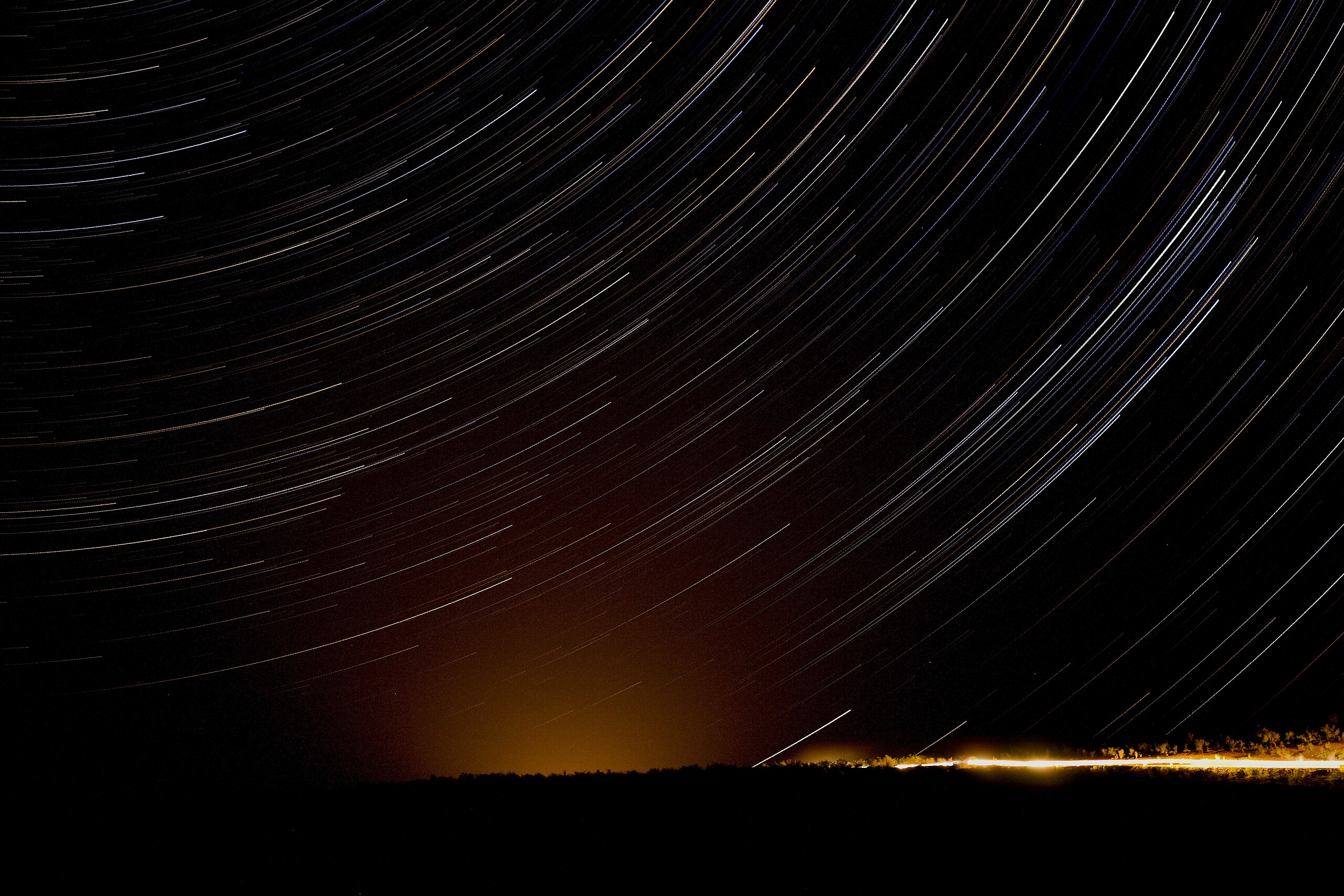 Star trails spin in circles in the sky over the Darvaza Crater, seen at the bottom right of the image at a bright light emanating from the ground.