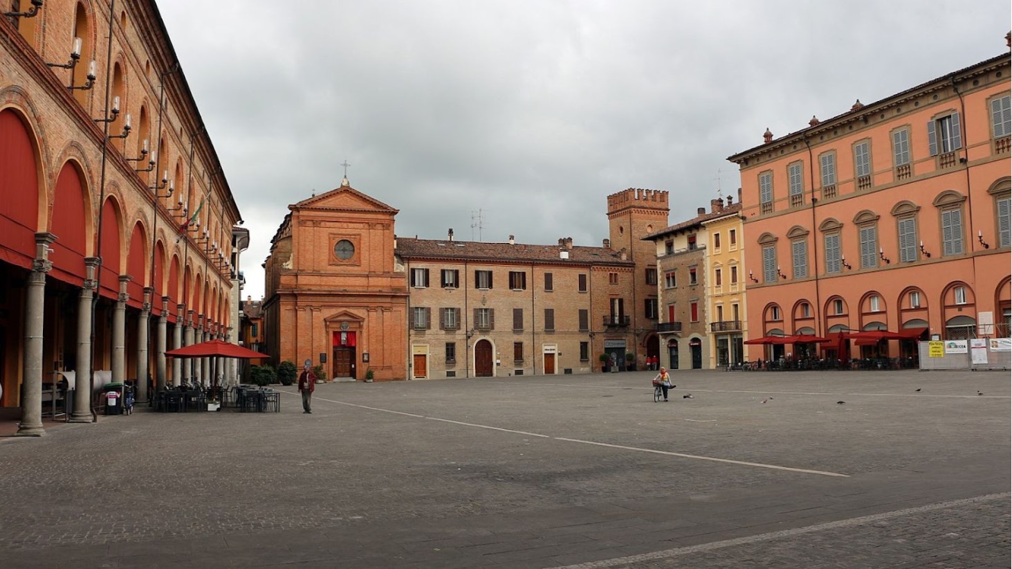 A spacious Italian piazza is captured in this image, surrounded by historic buildings with classical architectural features. On the left, a building with a row of arched porticos and on the right, a structure with a series of tall windows and balconies