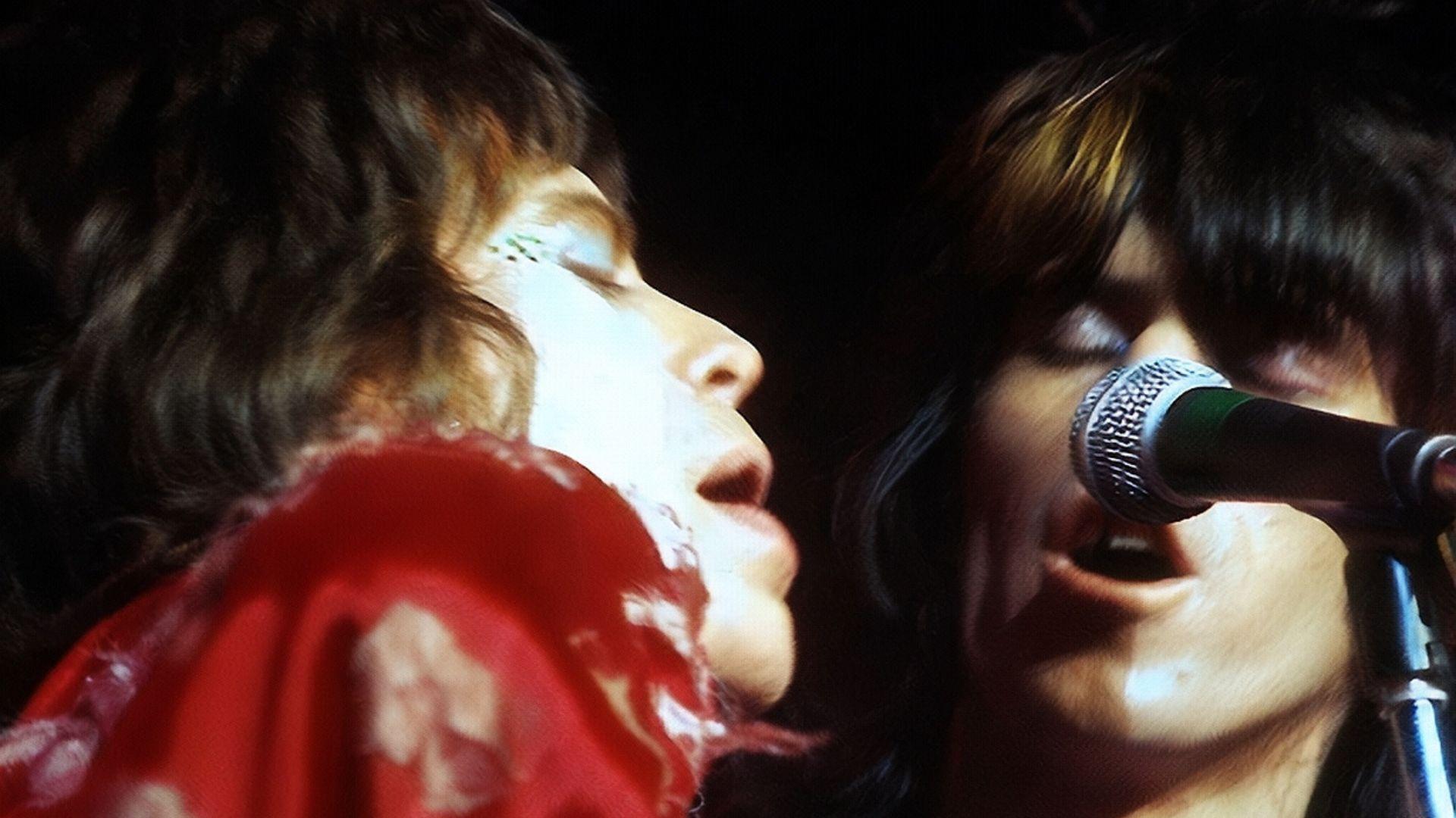 A close-up photo capturing Mick Jagger and Keith Richards sharing a microphone on stage. The image shows the singers in profile, with Jagger wearing a red scarf and both deeply engrossed in the performance