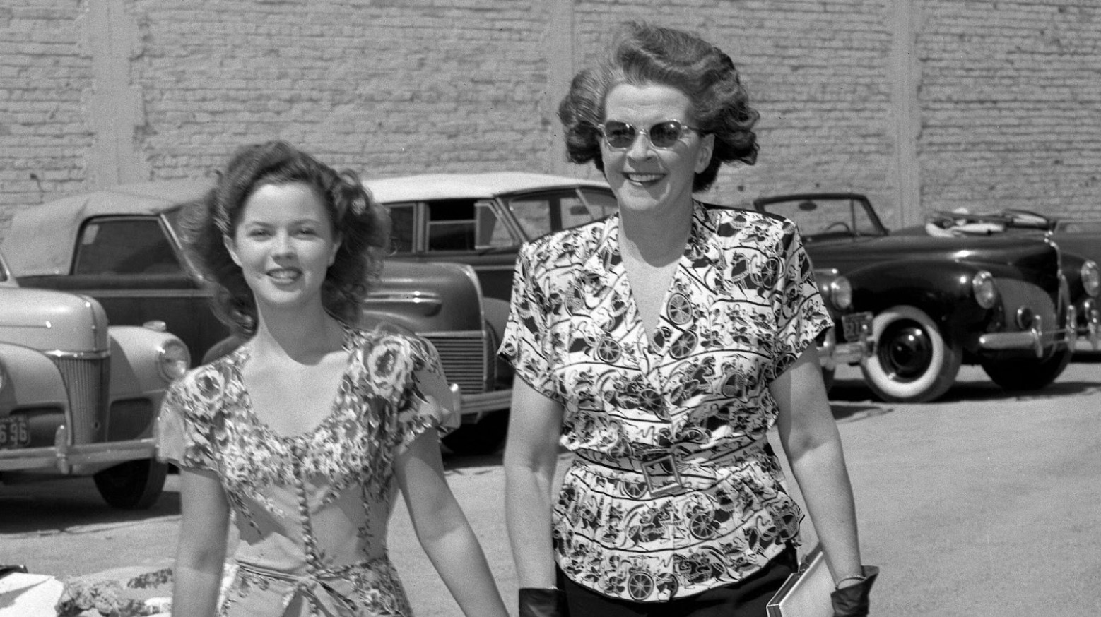 A black and white candid photo of Shirley Temple walking with her mother and holding her hand down the street, cars parked behind them, in 1945.