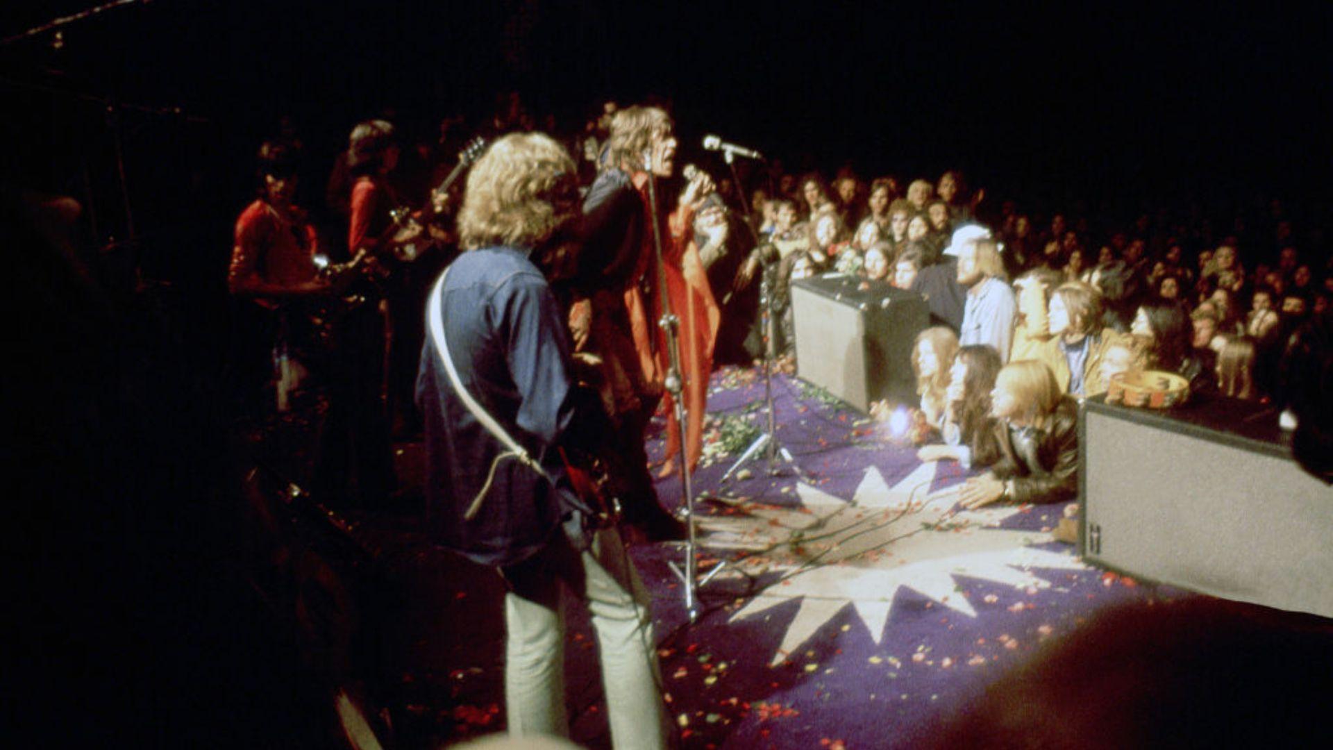 View from behind the Rolling Stones performing on stage at a rock concert, facing an audience. The band members are seen from the back, with one singer in a red shirt and scarf addressing the crowd at a microphone. The audience is in semi-darkness, with many faces looking towards the stage