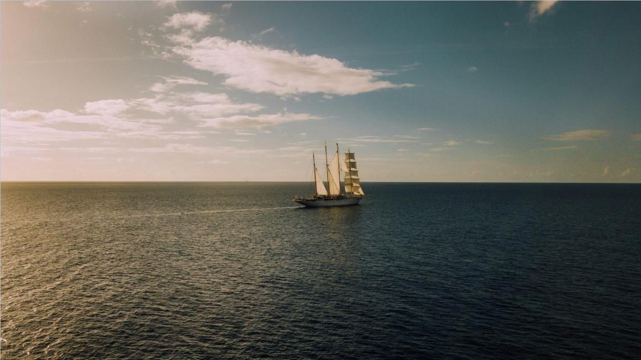 An old-looking ship on a body of water in the daytime under a blue and cloudy sky.