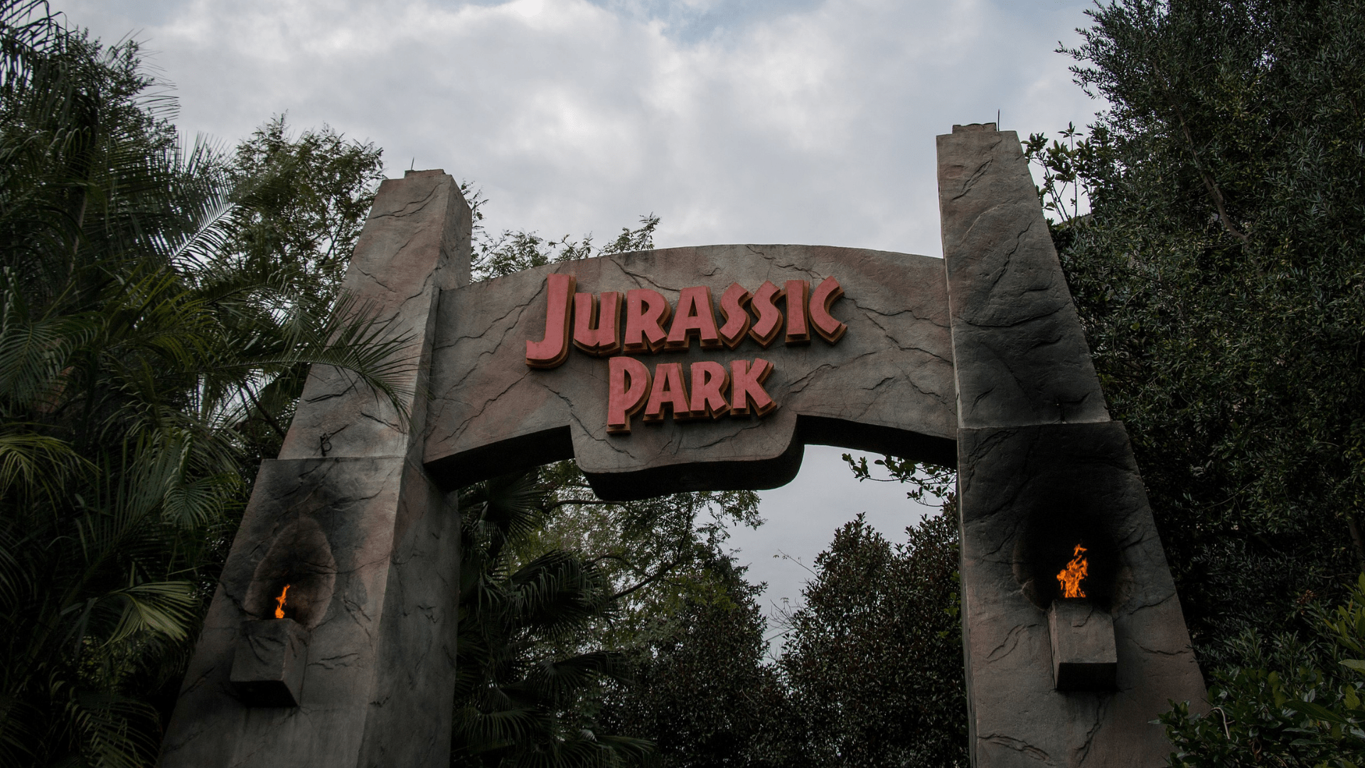 The entrance to the Jurassic Park ride at Universal Studios’ Islands of Adventure theme park. There is a large archway reading “Jurassic Park” with torches burning on either side, surrounded by tall trees and foliage.