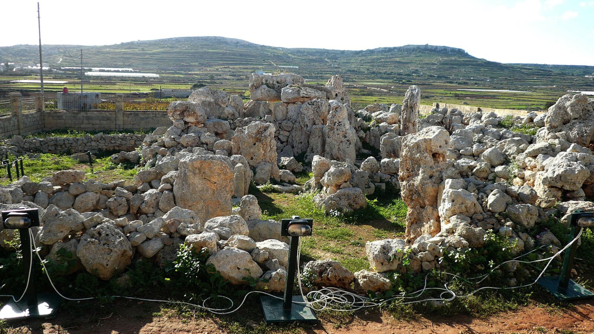 Image captures the sprawling ancient stone ruins in the Maltese countryside. The ruins consist of numerous large, weathered limestone blocks, some stacked to form walls and arches. In the background, terraced fields and a gentle hill characterize the rural landscape