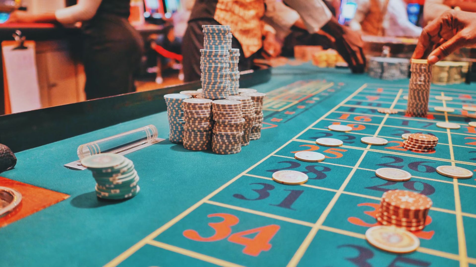 A table in a casino. There are people standing around it. The table is green with different numbers. There are some chips on the table. Some are piled up and others are on the numbers.