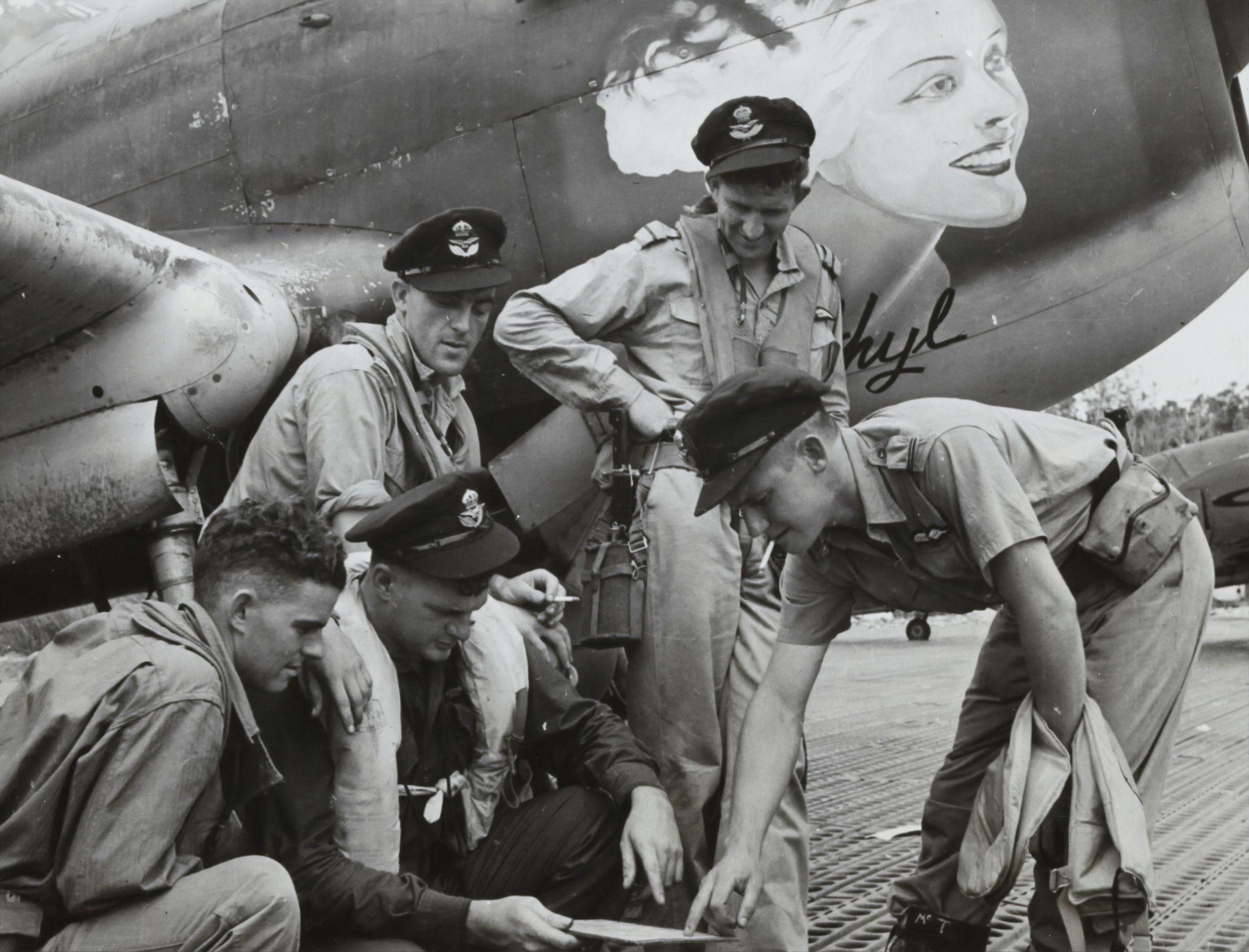 A squadron of pilots near a plane looking at a piece of paper during World War II.