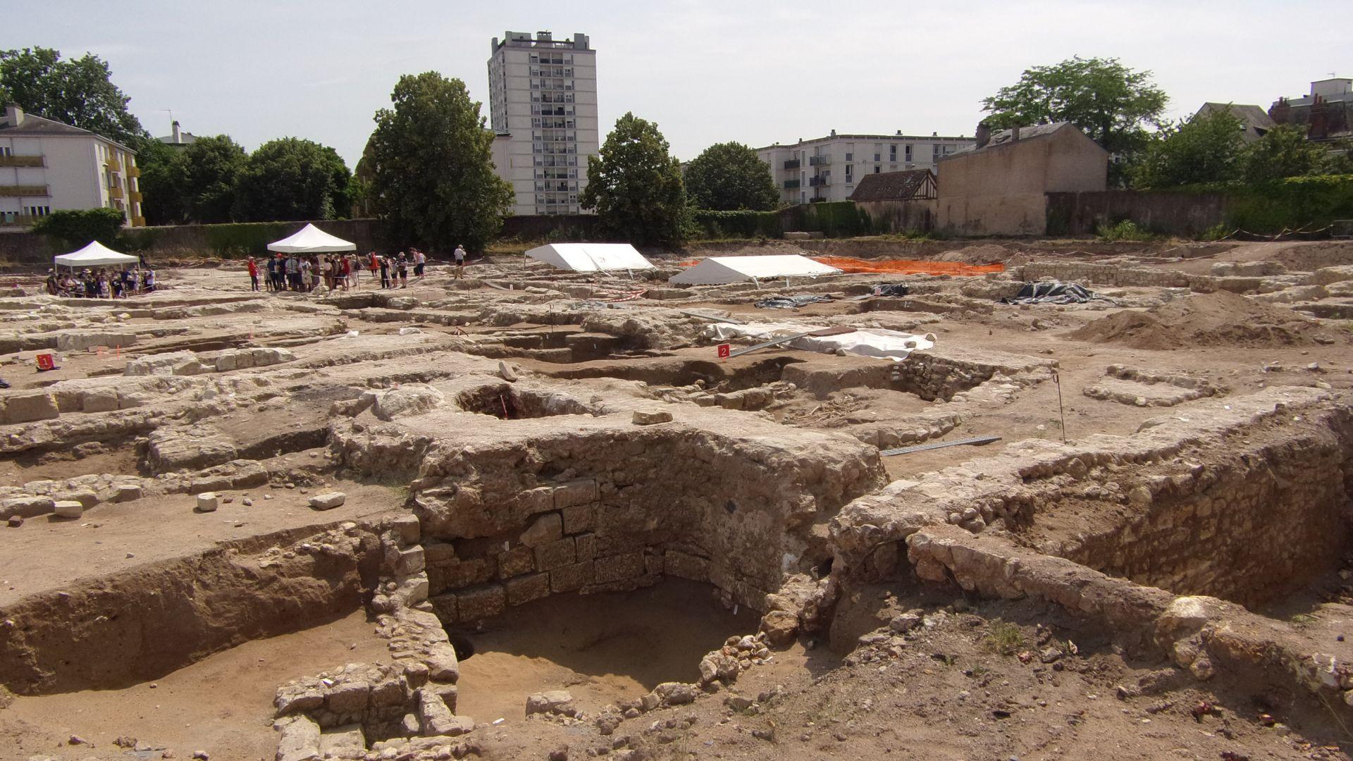 An area of the north part of Beaumont Abbey during an archeological dig, with modern-day buildings in the background.