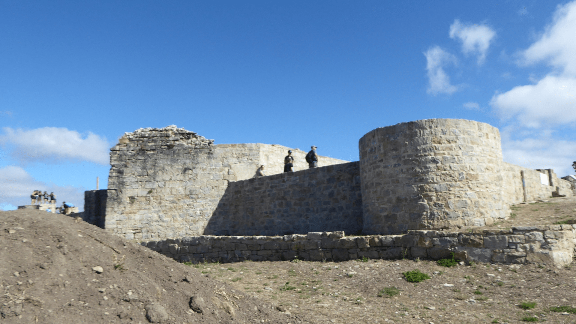 Some ruins at Irulegi in Spain. A group of people pose for a photo far in the background on the left of the image, while two people stand on top of a wall near the center of the image. The ruins look to be of an old fortification made of stone.