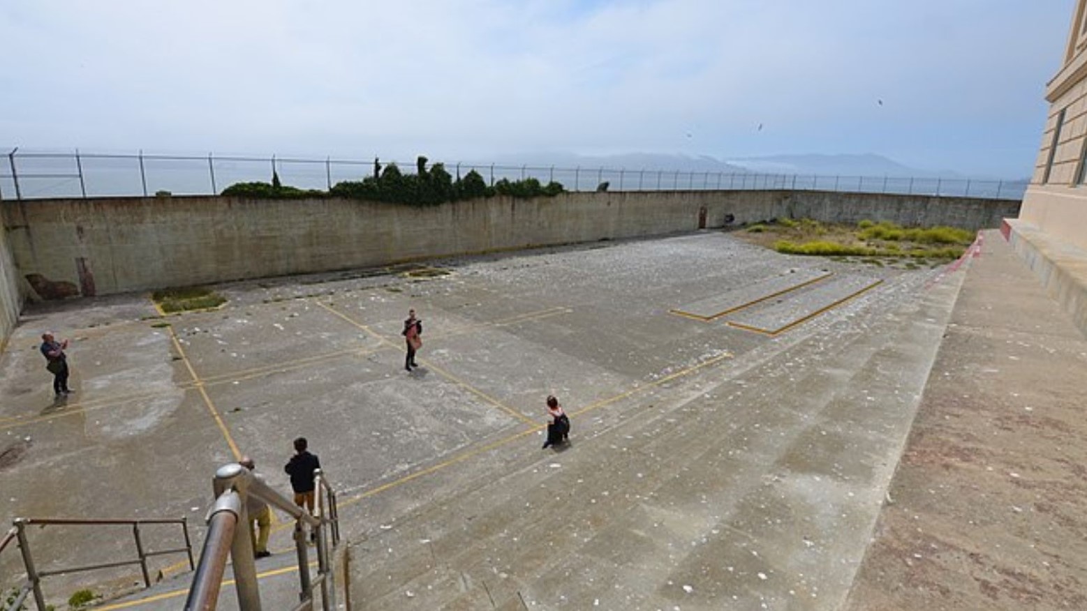 The Alcatraz prison recreation yard. The yard is made of concrete and some hills can be seen in the distance. There are some people in the yard and the high walls surrounding the yard make it hard to get out.