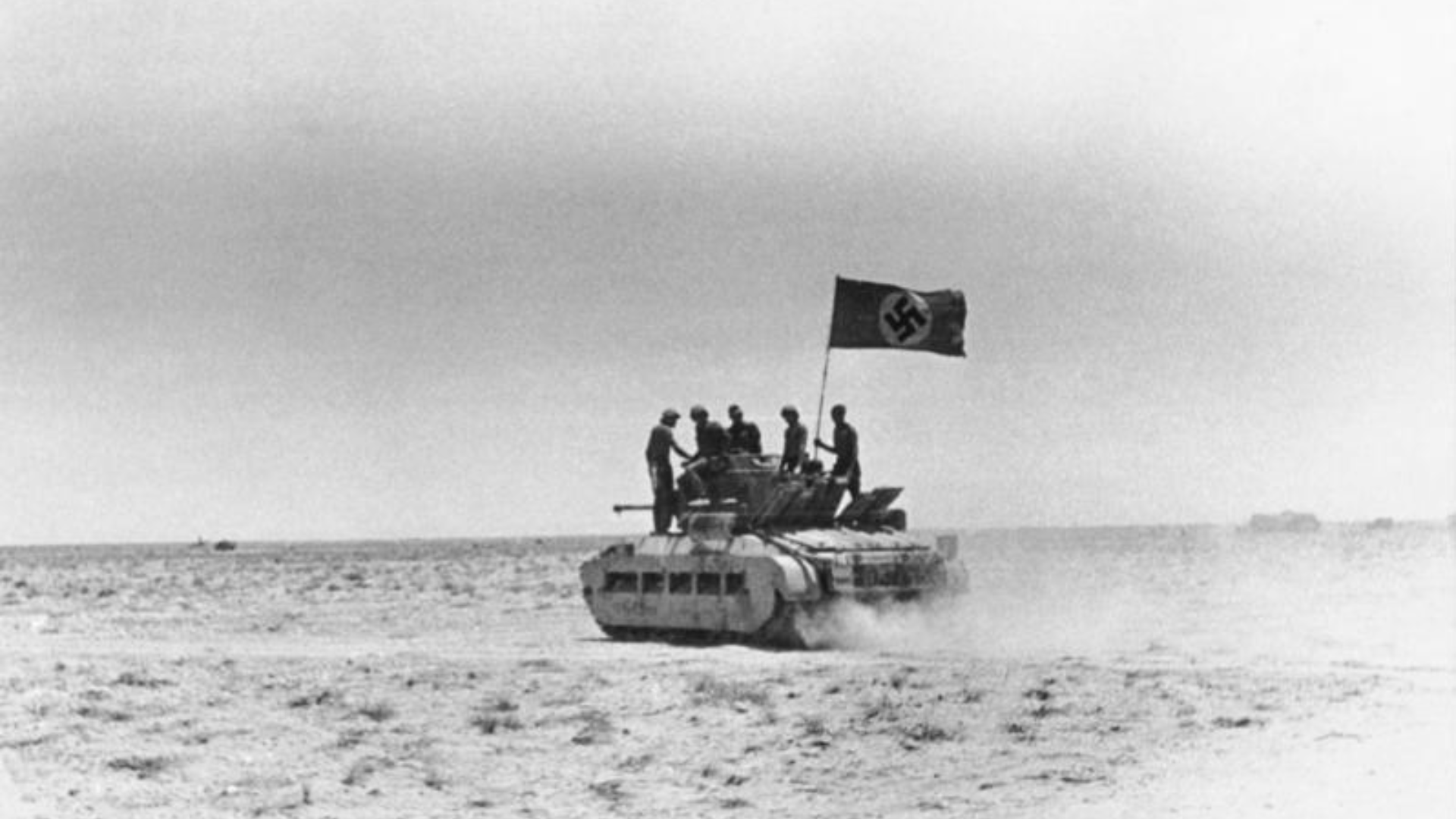 Black-and-white photo of an Afrika Korps Infantry-Panzer Mark II moving across the African desert with five men on top and a Nazi flag flying.