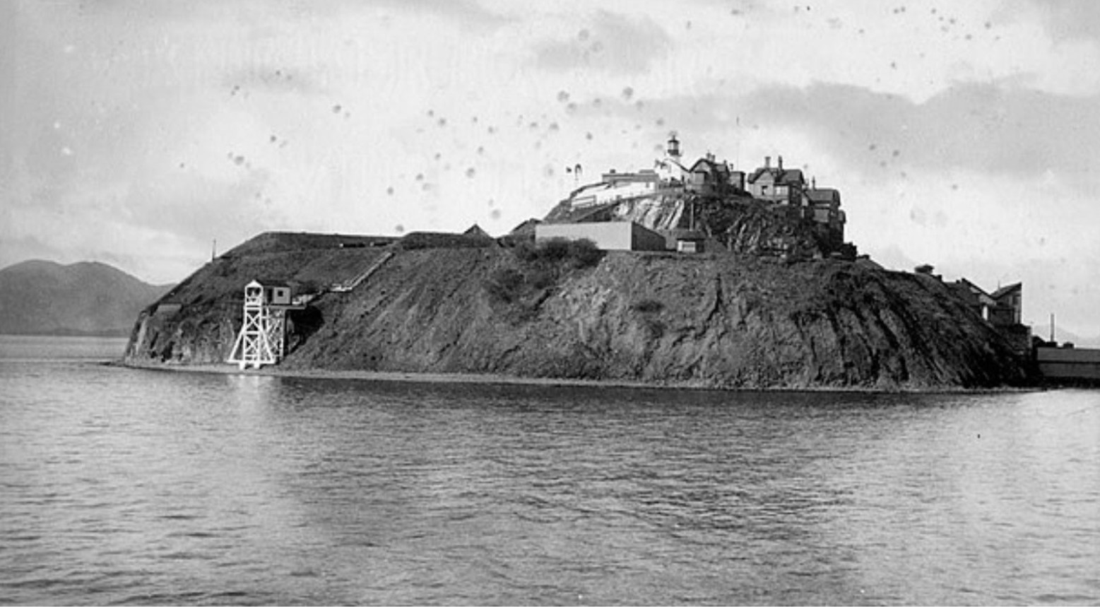 A black and white image of Alcatraz Island when it was a military base. The lighthouse is visible, as well as a few other houses at the top of the island.