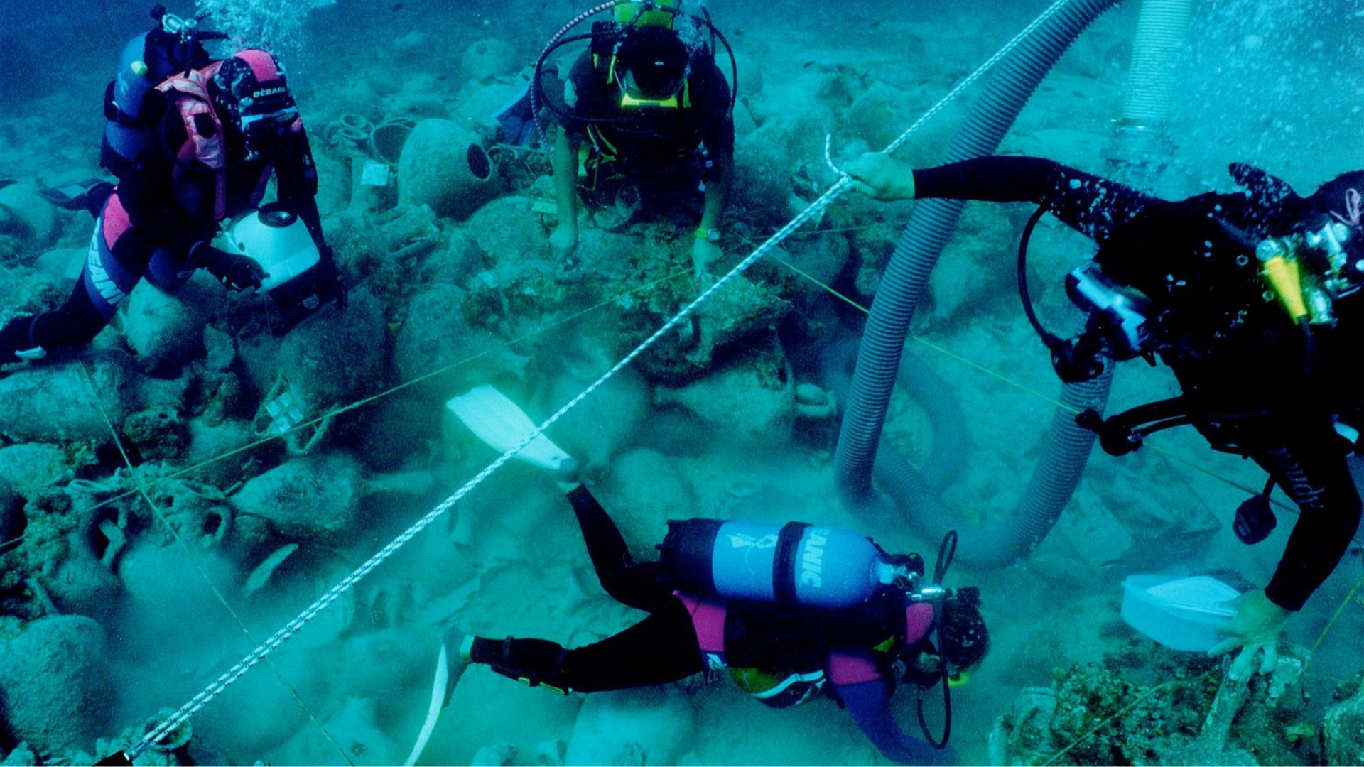 Underwater image showing three scuba divers engaged in an archaeological excavation. The divers, wearing full dive gear including fins, tanks, and masks, are submerged in blue-green water and are positioned around a site marked with measuring lines. Visible on the sea floor among the divers are various ancient amphorae and scattered stones