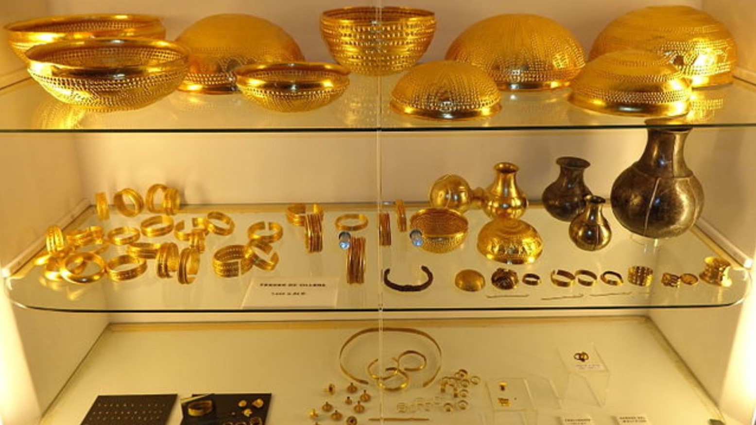 The Treasure of Villena on shelves. The top shelf has gold bowls, the middle has gold bracelets and vases, and the bottom shelf has some more gold bracelets and rings.