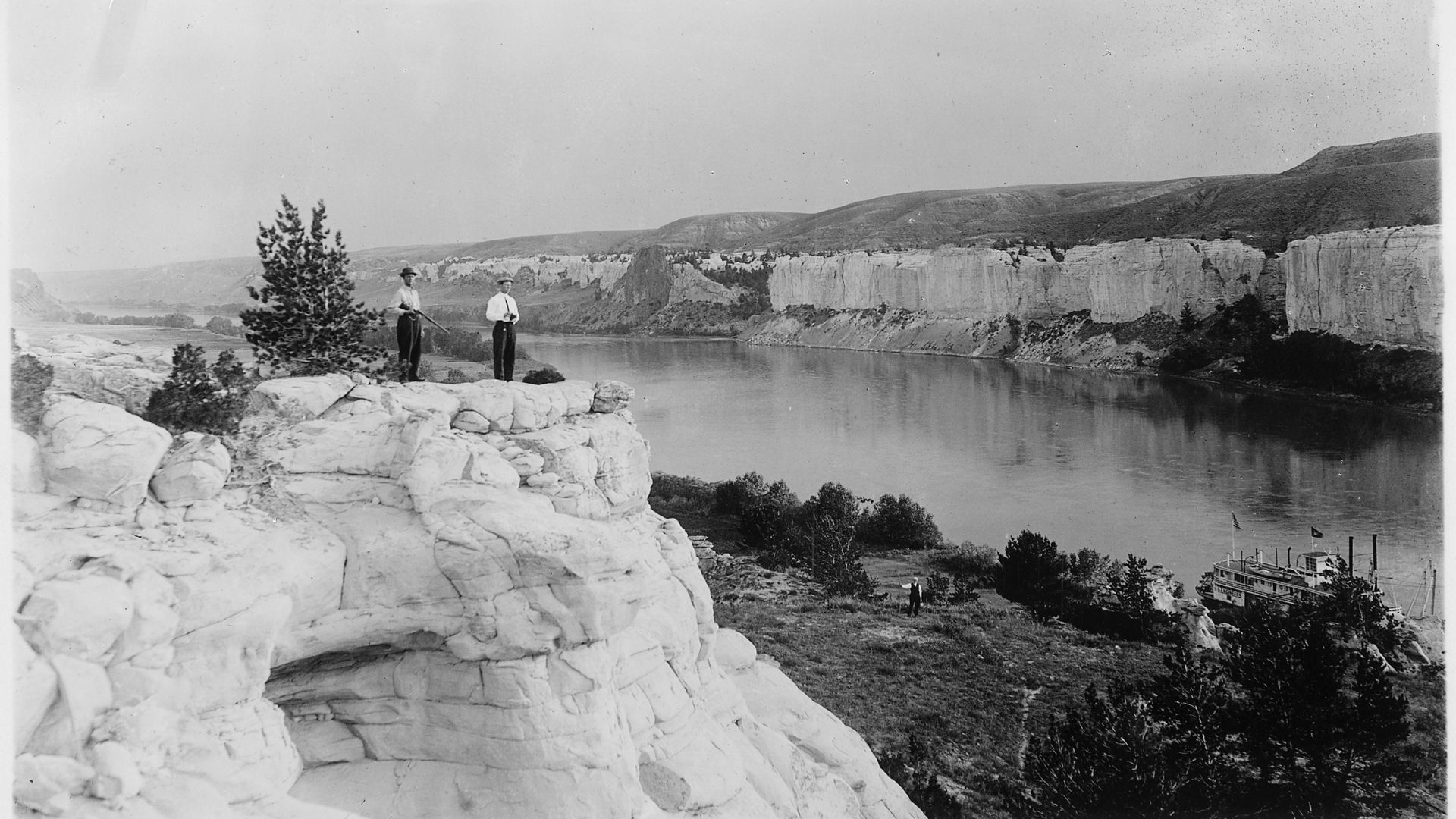 Two men standing on a cliff overlooking the Missouri River that holds a steamboat in a 1912 photo.