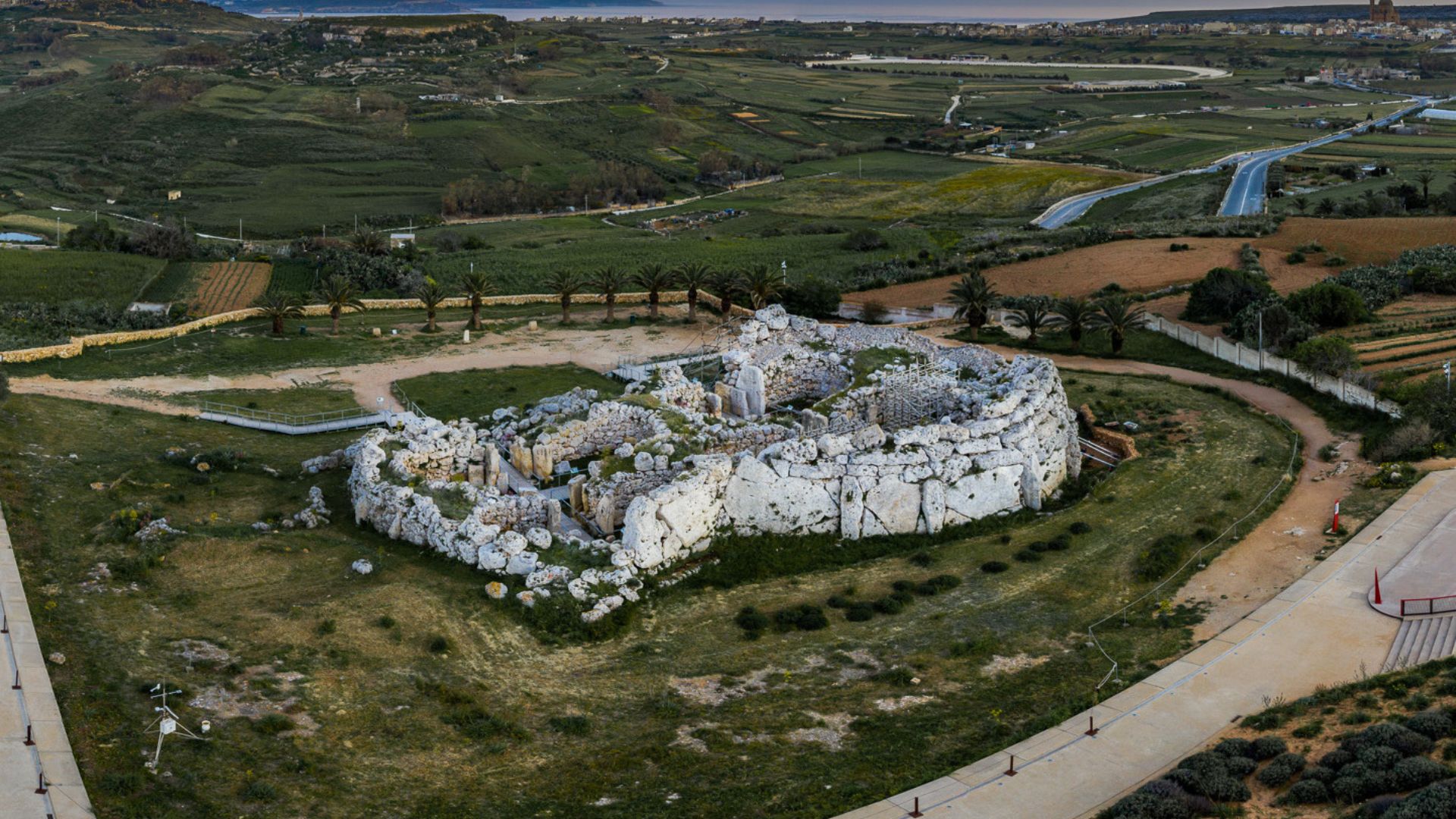 An aerial view of the ancient megalithic temple ruins located in the Maltese countryside. The ruins are comprised of large white limestone blocks forming a semi-circular structure surrounded by pathways and sparse vegetation