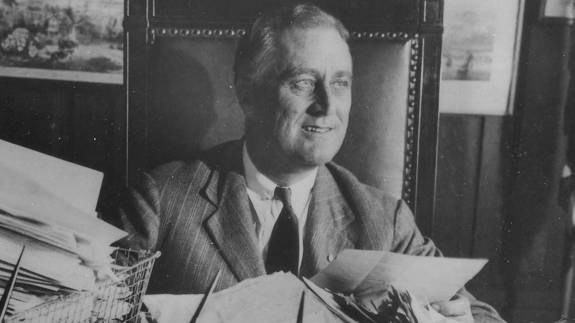 Black-and-white photo of Franklin D. Roosevelt sits at a desk holding a document, wearing a suit, and smiling.