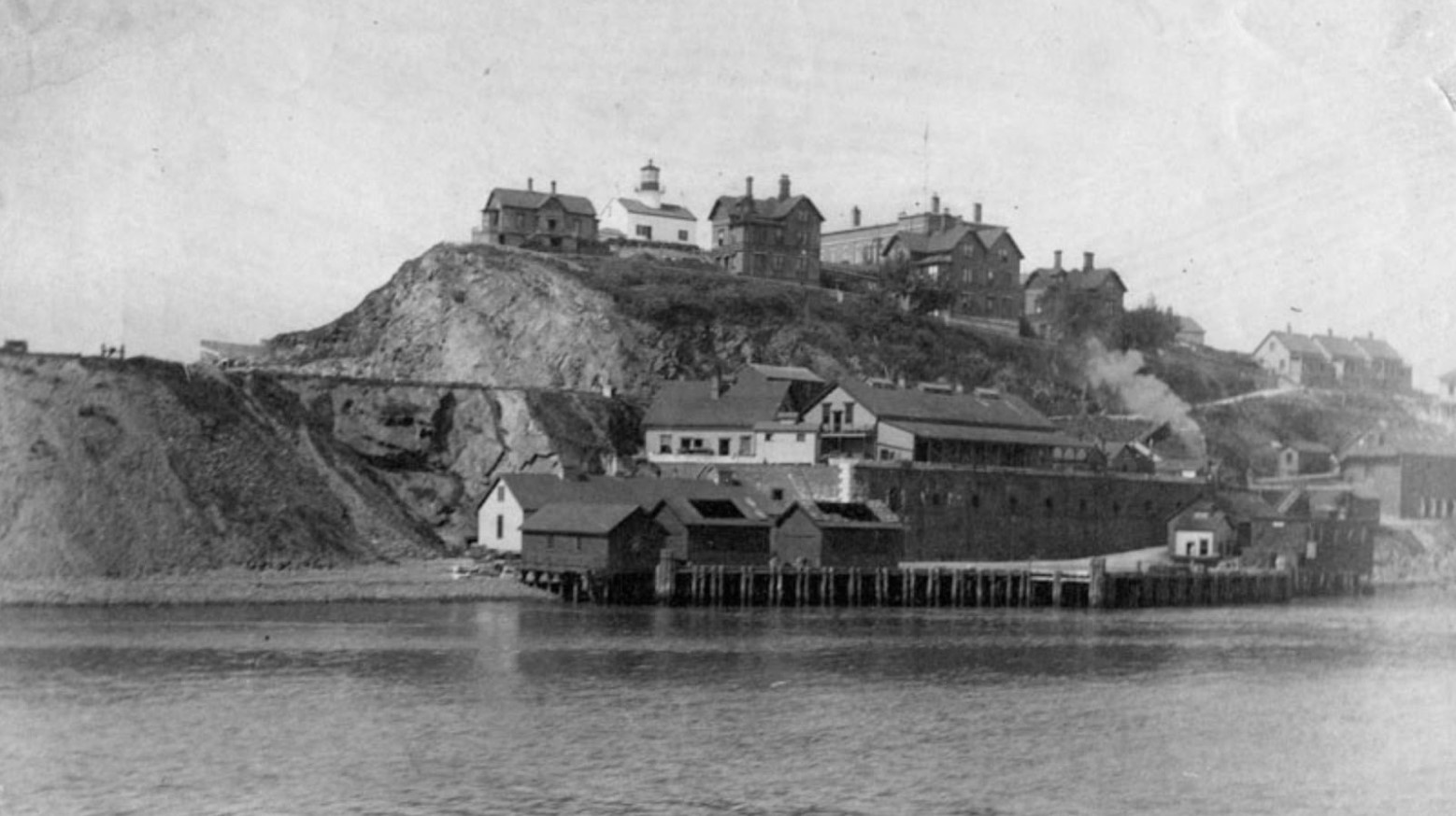 A black and white image of Alcatraz Island when it was a military base. There is a port at the bottom of the island and various houses up on the hill.