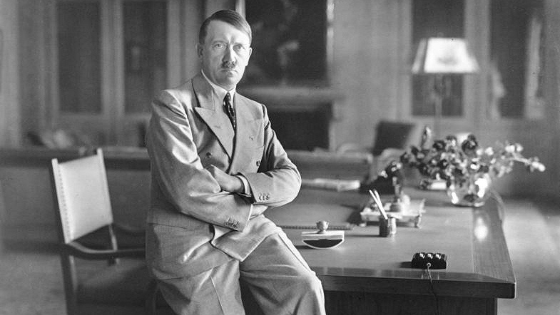 Black-and-white photo of Adolf Hitler wearing a suit and sitting on the edge of his desk prior to World War II.