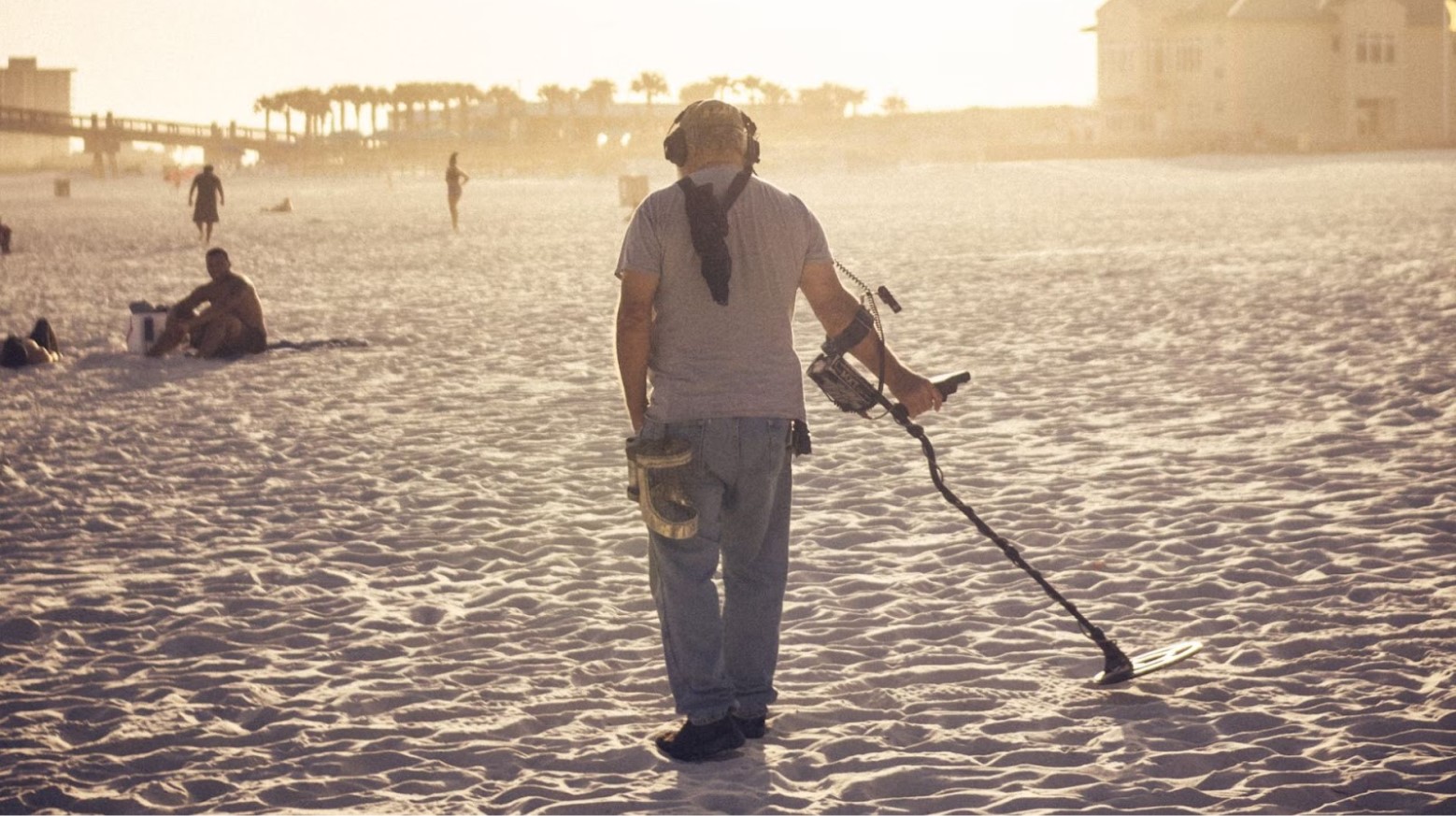 A man on a beach with a metal detector searching for treasure. Some other people are sitting on the beach and playing in the sand. There are some palm trees and buildings in the distance.