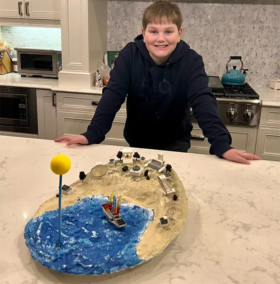 A young man in a black hoodie standing at a kitchen counter with his model of the ancient death ray