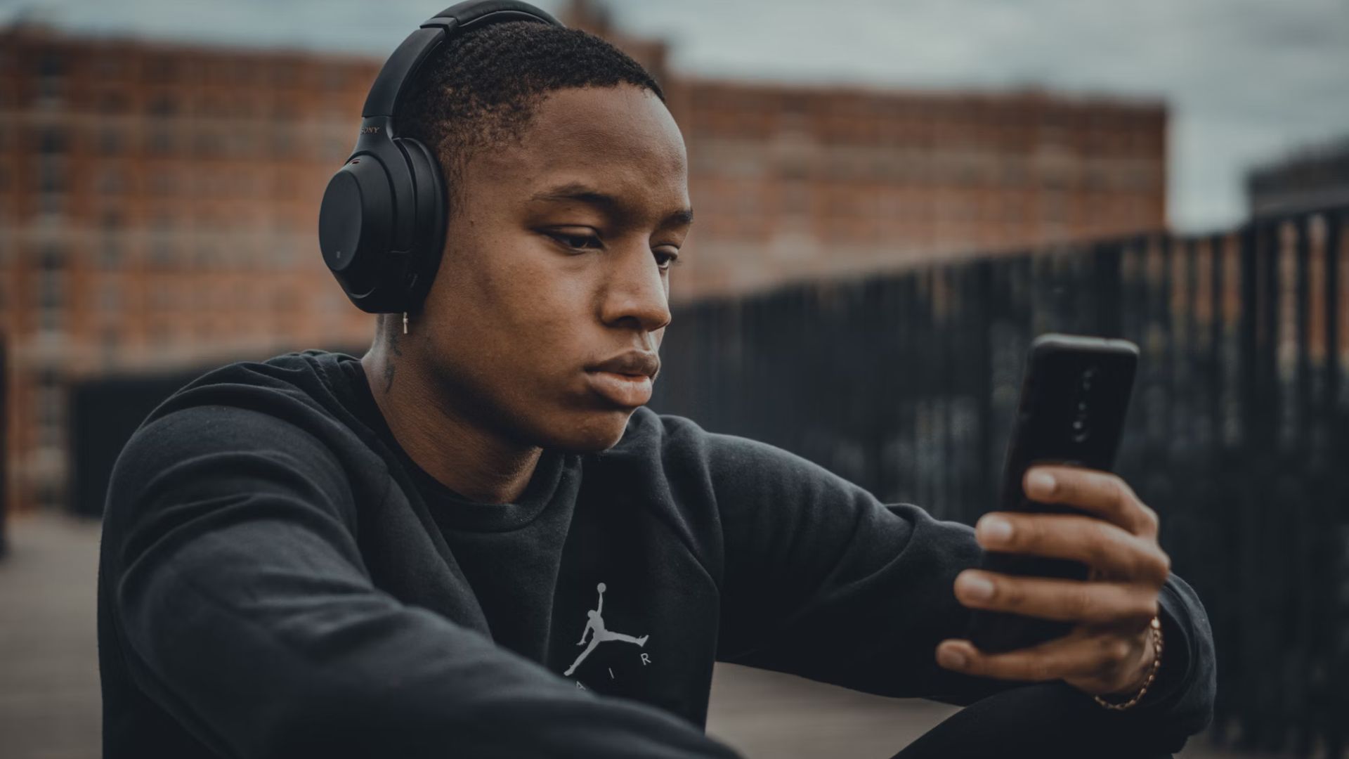 A man sitting outside listening to music. He is wearing a black jumper, black headphones, and is holding a black phone.